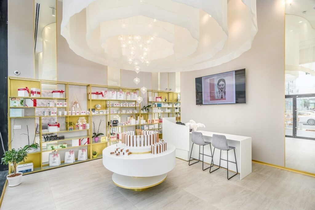 זוהי תמונה של חנות קוסמטיקה יוקרתית עם מדפים מלאים במוצרים, שולחן תצוגה עגול ודלפק קבלה עם מסך טלווי