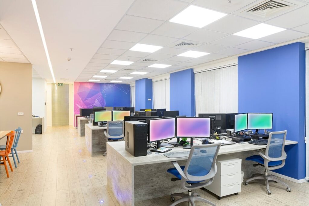 משרד מרווח עם שורות של שולחנות עבודה, מחשבים, וכיסאות, קירות בצבע כחול ותקרה מוארת היטב.