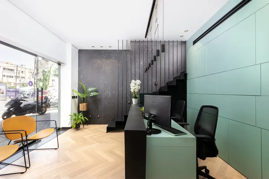 תמונה של חלל פנימי מודרני עם דלפק קבלה ירוק בהיר, מדרגות שחורות, רצפת עץ, וחלון גדול שמציג רח