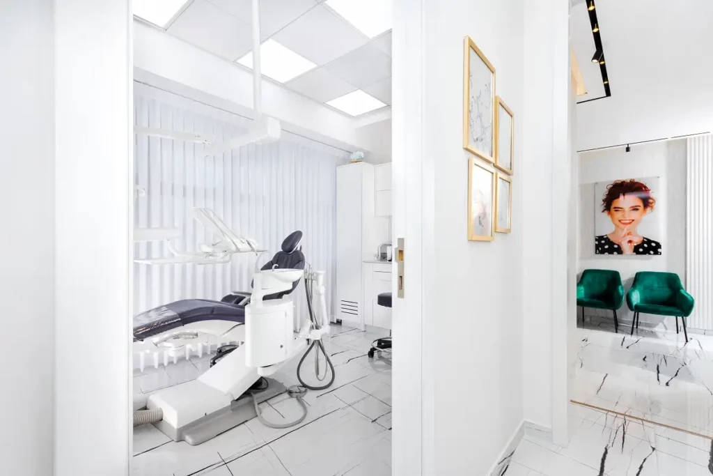 מבט לתוך מרפאת שיניים מודרנית ובהירה, עם כיסא טיפולים כחול-אפור, וחדר המתנה עם תמונות וכיסאות