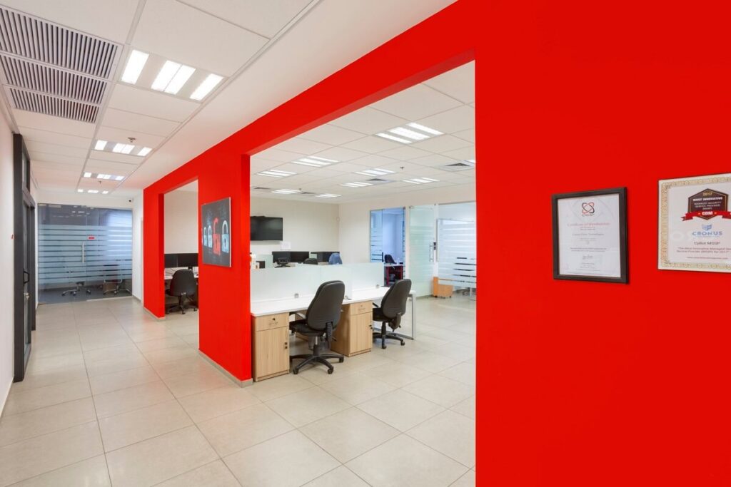 משרד מודרני עם קירות אדומים, עמדות עבודה מסודרות, ותעודות תלויות על הקיר.