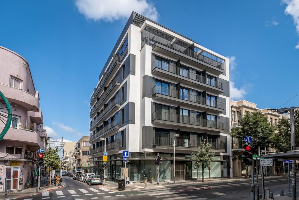 בצילום רחוב בעיר, בניין מגורים מודרני בן שש קומות עם מרפסות שחורות ניצב לצד בניין ישן יותר, וברקע שמיים כ