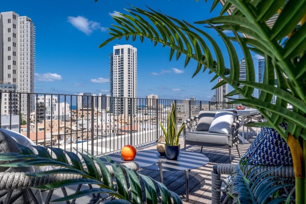 מרפסת גג עם ריהוט גן, צמחייה ועיר ברקע תחת שמיים כחולים.