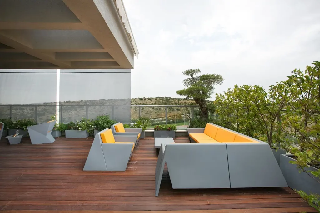 מרפסת עץ עם ריהוט גן אפור וכריות כתומות, צמחייה ונוף הררי ברקע.