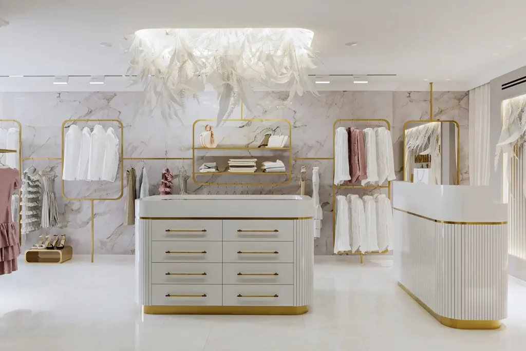 בחדר בהיר ומרווח מוצגים בגדים על קולבים מזהב, לצד שידה לבנה עם מגירות ודלפק לבן, ועל התקרה תלויה נברשת נוצות