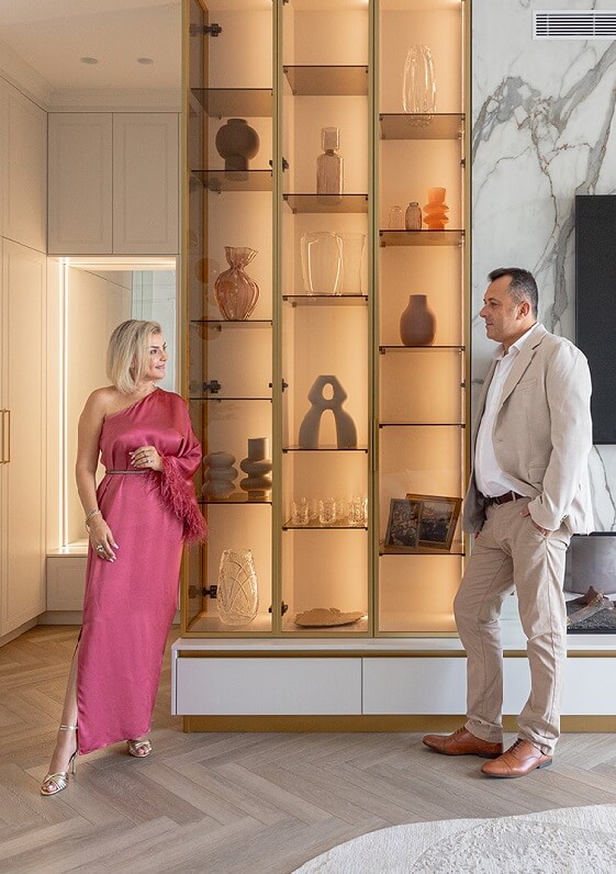 אישה בשמלה ורודה וגבר בחליפה בהירה עומדים בחדר מואר ליד ארון תצוגה עם כלי נוי.