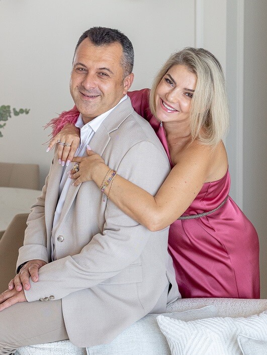 בתמונה גבר בחליפה בהירה ואישה בשמלה ורודה מחובקים וחייכניים, יושבים על ספה.